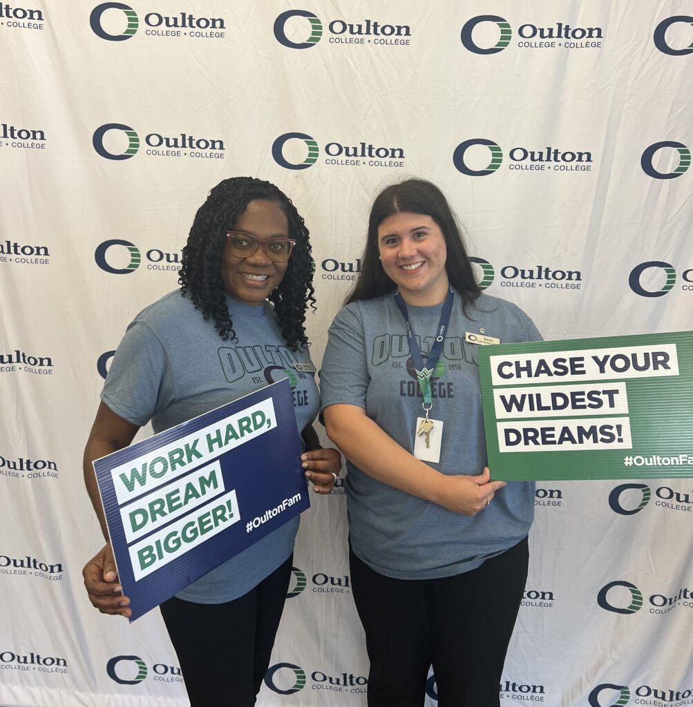 Two Oulton College employees smiling in front of an Oulton College backdrop, holding signs with inspirational words.