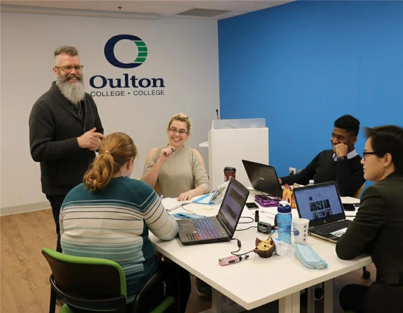 Business administration students at Oulton College. Four students stitting around a table with an instructor teaching Business administration and Business managemnet concepts, as part of their Associates Degree in Business Administration.