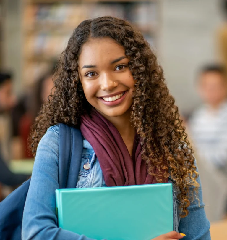 Female student, holding some files in her hand, wearing a purple scarf and Blue shirt. She is smiling and looking forward to starting her Business Management & Entrepreneurship Program at Oulton College.